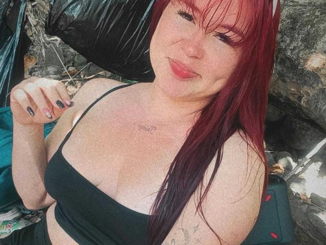 KendraVelvet with red hair, black tank top, and tattoos, pouting with her tongue out, showing her tits, and holding her finger to her cheek. Her nails are painted black.
