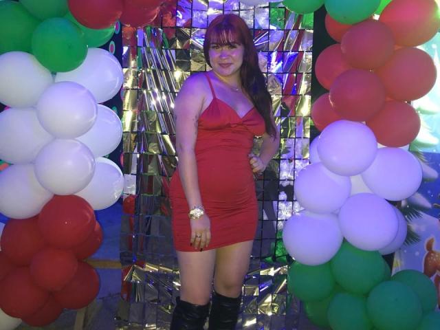 KendraVelvet stands in a red dress, showing her tits, with a smile, hands on hips, surrounded by balloons, wearing black boots, against a shiny, metallic background.