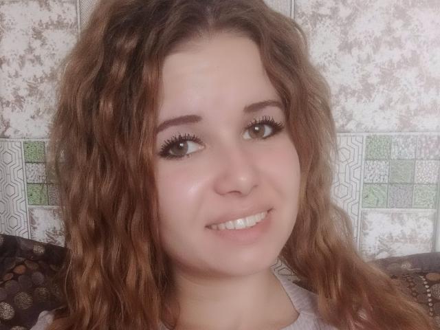 BertieMyra has curly brown hair, light skin, and is smiling with long eyelashes, showing off her white teeth. She's wearing a grey top. Her tits are not visible.