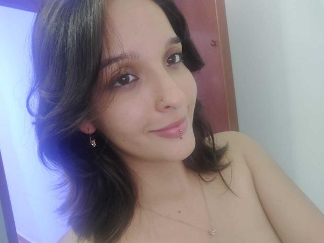 LanaKlaire, con cabello castaño de largo medio, perforación en la nariz, perforación en el labio, brillo labial rosa, aretes pequeños, collar, sonriendo, sin top, mostrando los hombros desnudos..