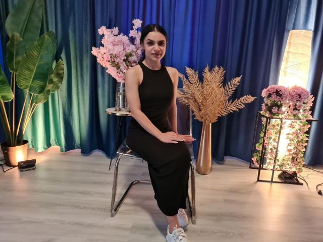 MiniMira with dark hair, black sleeveless dress, and white sneakers, sitting on a chrome chair, smiling, with pink flowers and blue curtains in the background.