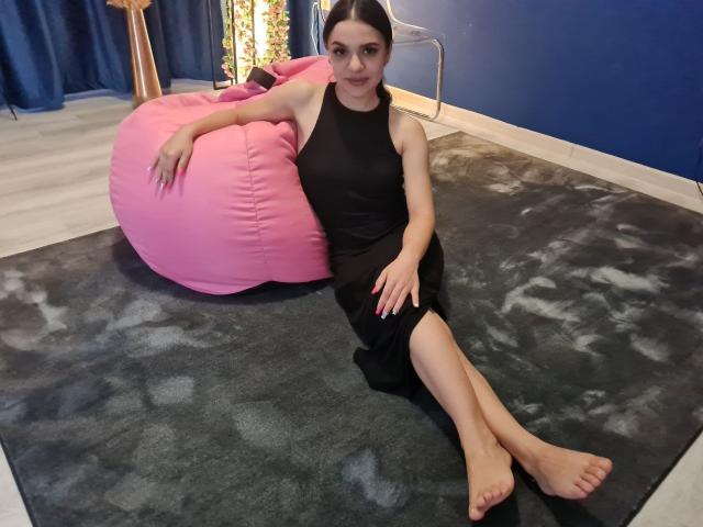 MiniMira with bare feet, black dress, and pink beanbag, sitting on a grey rug, arms resting, looking at camera, dark hair, blue wall in background.
