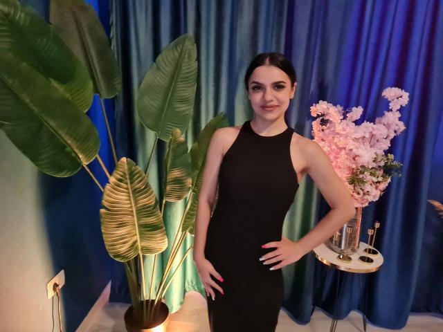MiniMira stands in a black dress, hands on hips, smiling with dark hair in a ponytail. She's flanked by green plants and pink flowers on a white table.