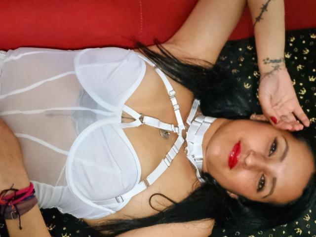 XimenaNahomi is lying on a red couch, wearing a sheer white lingerie set with straps, exposing her tits. She has red lipstick, black hair, and a tattoo on her arm.