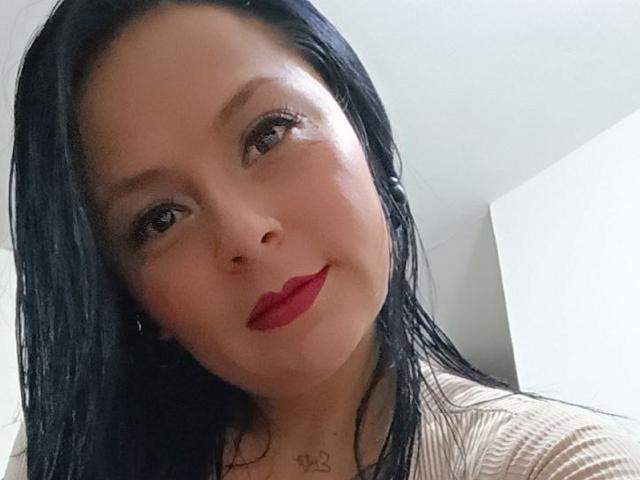 XimenaNahomi has black hair, red lipstick, and a subtle smile. She's wearing a beige top with a pattern, and her chest has a small tattoo.