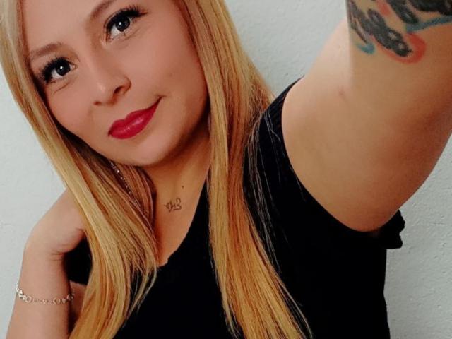 XimenaNahomi has blonde hair, red lipstick, black shirt, tattoo on arm, and bracelet. She's smiling with a cute face, showing her tits slightly with a raised arm.