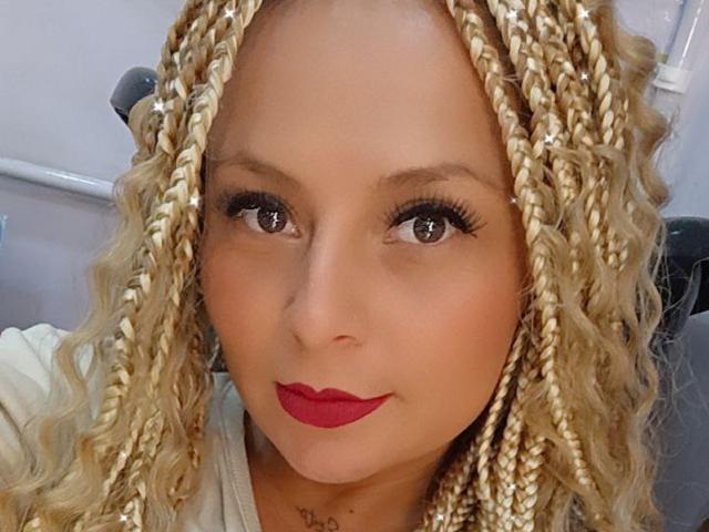XimenaNahomi has blonde braids, red lipstick, and dark eyeliner. Her brown eyes are slightly upturned, and she's smiling softly at the camera. She's wearing a white shirt.