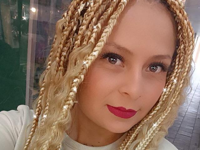 XimenaNahomi has blonde braids, red lipstick, and brown eyes, wearing a white top, with a slight smile, in a selfie. Her tits are not visible.