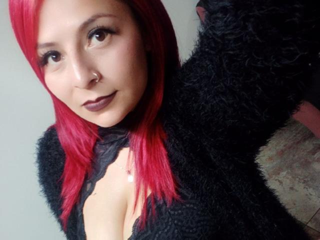 XimenaNahomi has red hair, dark makeup, a nose ring, and is wearing a black fuzzy top with a visible cleavage, looking at the camera with a slight smile.