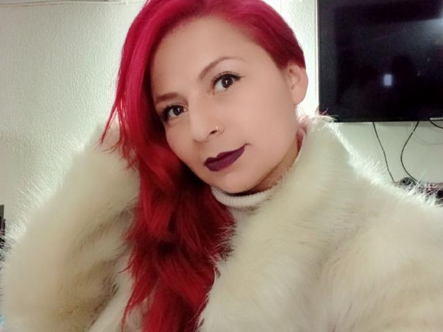 XimenaNahomi with red hair, white fur coat, and purple lipstick, smiling at the camera. She has a nose ring and dark eye makeup. Her tits are partially covered.
