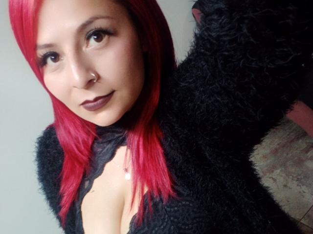 XimenaNahomi has red hair, dark lipstick, a nose ring, and wears a black fuzzy top with cleavage, looking at the camera with a slight smile.