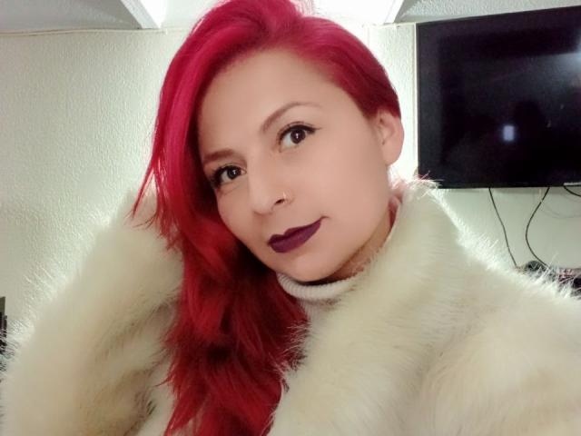 XimenaNahomi with bright red hair, wearing a fluffy white coat, dark purple lipstick, and a slight smile, looking at the camera with a relaxed, confident expression.