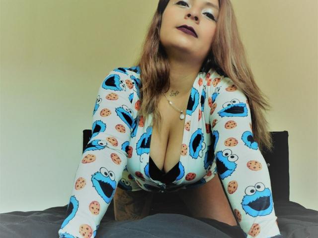 XimenaNahomi with big tits, wearing a blue Cookie Monster pajama top, black bra, and purple lipstick, leaning forward on a bed, looking up with a sultry expression.