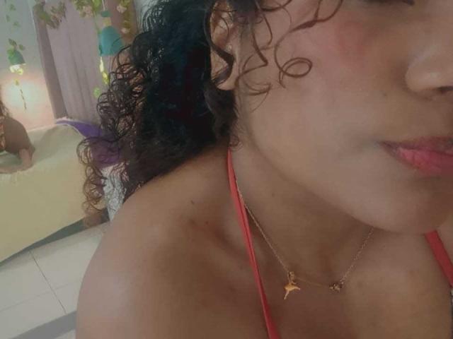 EvaBennet with curly brown hair, red halter top, pink lips, slight blush, gold necklace with small animal pendant, visible shoulder, soft lighting, slightly turned head.