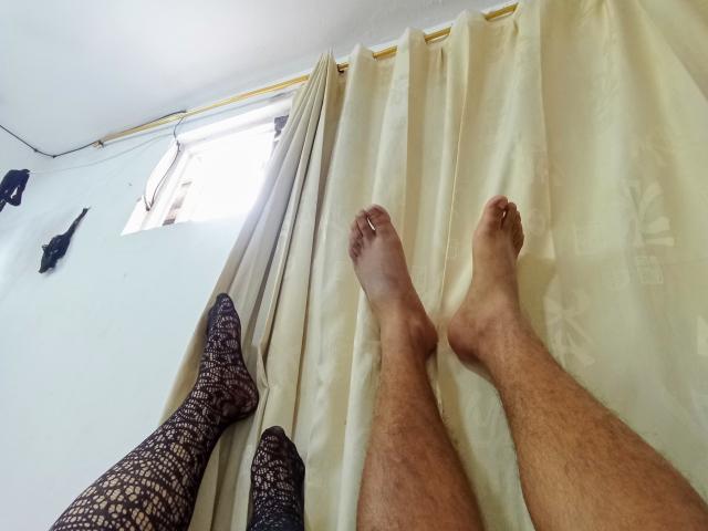 LatinStyleX's legs, wearing black lace stockings, with bare feet, resting against beige curtains in a room with a small window and white walls.