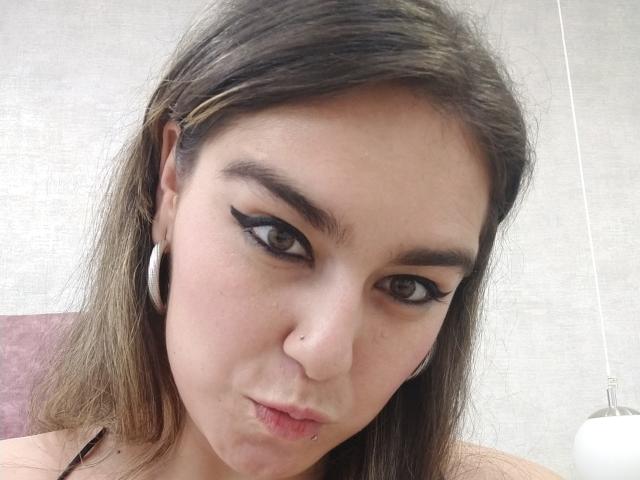 TaylorNova, avec des lèvres charnues, des yeux marron, un trait d'eye-liner noir, un piercing au nez, de petites boucles d'oreilles et des cheveux bruns lisses, regarde directement l'appareil photo. Son visage est légèrement rougi..