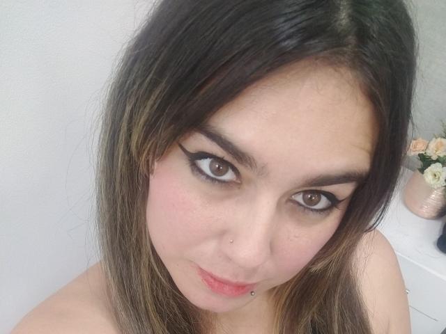 A TaylorNova tem olhos castanhos, delineador preto e uma pequena perfuração no nariz. Ela está olhando para cima com um leve sorriso, e seu cabelo castanho cai ao redor do rosto. Seus seios não estão visíveis..