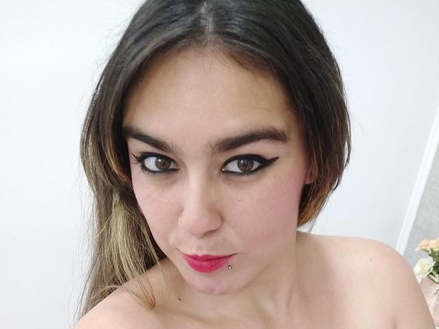 TaylorNova, avec ses yeux marron foncé, son eye-liner noir, son rouge à lèvres rose et son piercing sur la lèvre inférieure, a de longs cheveux bruns et une expression légèrement boudeuse, mettant en valeur ses épaules dénudées..