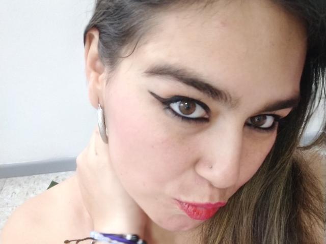 TaylorNova's face with brown eyes, black eyeliner, pink lips, and a small nose ring. She has light skin, dark brown hair, and wears hoop earrings. Her boobs are barely visible.