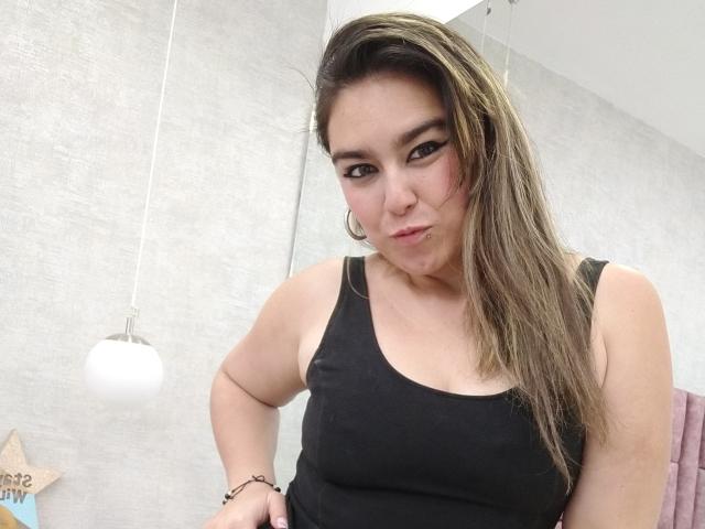 TaylorNova, com cabelo castanho claro, camiseta preta, brincos grandes e piercing no nariz, fazendo um leve biquinho, olhando para a câmera, com as mãos nos quadris, em um banheiro com uma luminária pendente..