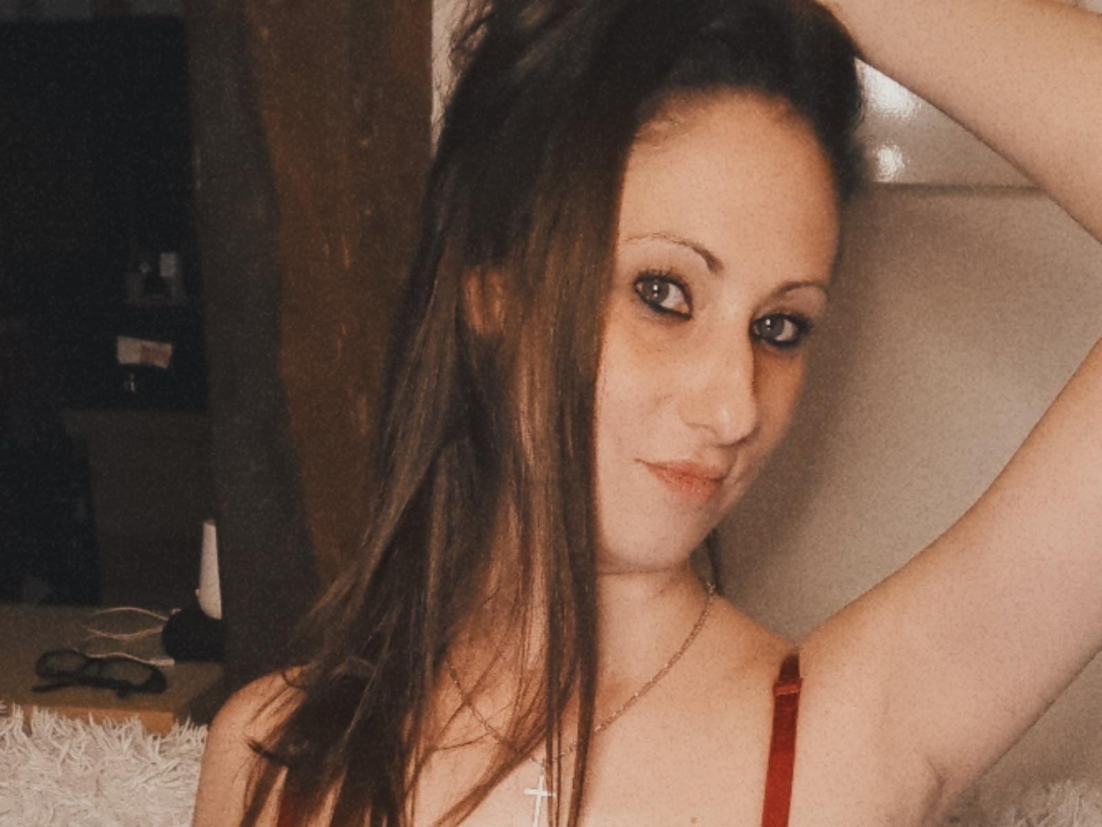 LynaThalion with long brown hair, arms raised, wearing a red strap, smiling slightly, small nose, blue eyes, pale skin, necklace, soft lighting, subtle makeup, casual selfie.