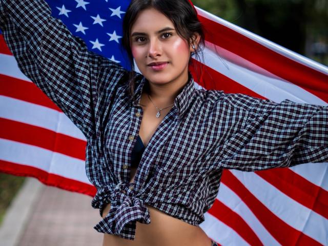 AnnieHotStar with tied plaid shirt, exposing black bra, holding up American flag, arms spread, smiling, dark hair, medium breasts, cute, outdoor, daytime.