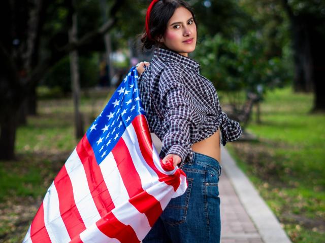 AnnieHotStar with a red bandana, checking over her shoulder, holding an American flag, wearing a tied check shirt, and blue jeans, standing in a grassy park.