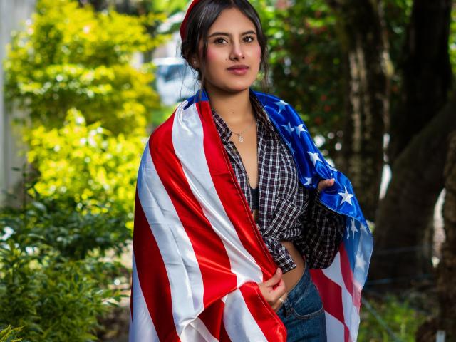 AnnieHotStar with an American flag draped over her shoulders, wearing a checkered shirt and jeans, standing outdoors, looking straight at the camera, with a neutral expression.