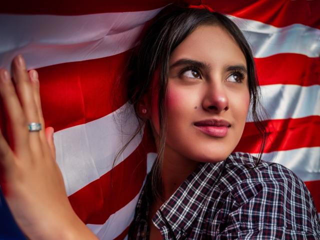 AnnieHotStar with brown eyes, dark hair in a ponytail, wearing a plaid shirt, touching a flag, has a cute smile, small mole on her right cheek, ring on her left hand.