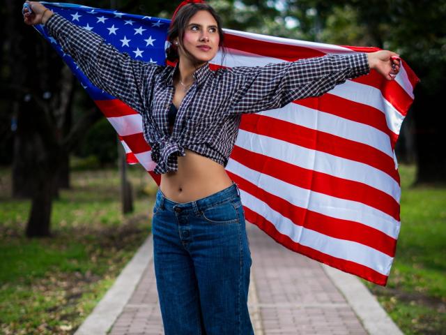 AnnieHotStar with a checkered shirt tied above her boobs, holding a large American flag, standing on a brick path, wearing blue jeans, looking up with a slight smile.