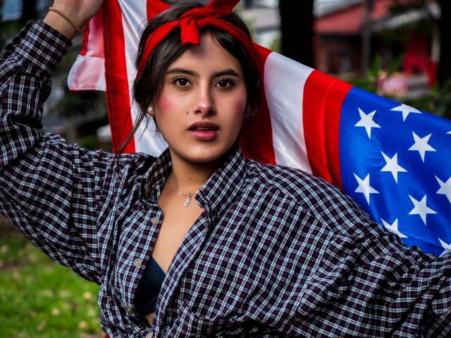 AnnieHotStar with red headband, plaid shirt, and black bra, holding an American flag, has a serious face, dark hair, and a small necklace. Background is blurred.