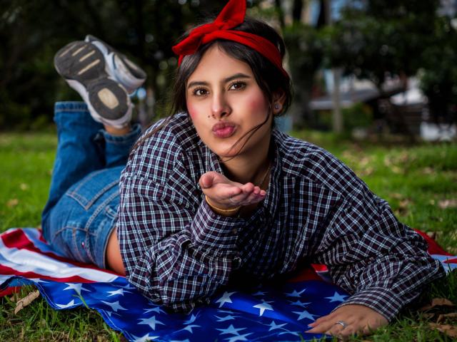 AnnieHotStar, lying on a star-spangled blanket, wearing a red headband, plaid shirt, and jeans, puffs her pink lips, showing a ring, with her feet up.