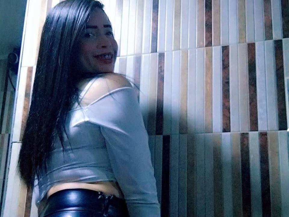 ValentinaLife with long black hair, smiling, wearing a white off-shoulder top and black leather pants, looking back over her shoulder. Boobs visible through top.