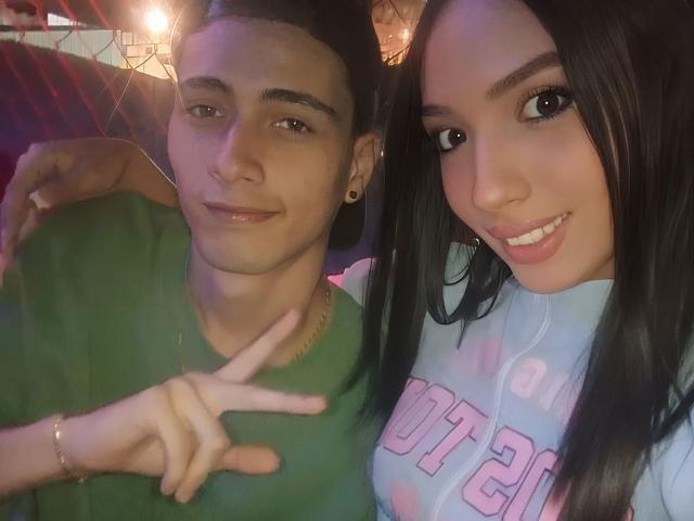 MarkusAndHenderly with short dark hair, green shirt, ear gauges, and a gold chain, making a peace sign, next to Henderly with long black hair, blue shirt, and pink text.