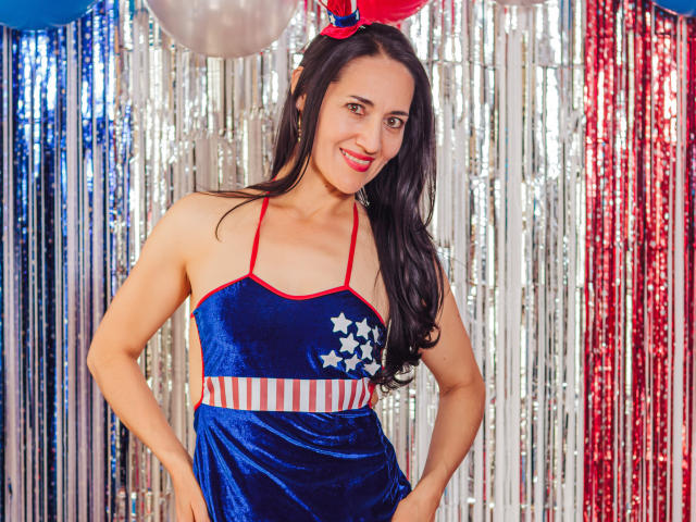 MiaMott with long black hair, wearing a blue velvet dress with red straps, white stars, and red-white stripes, smiling, hands on hips, red and silver bunny ears.