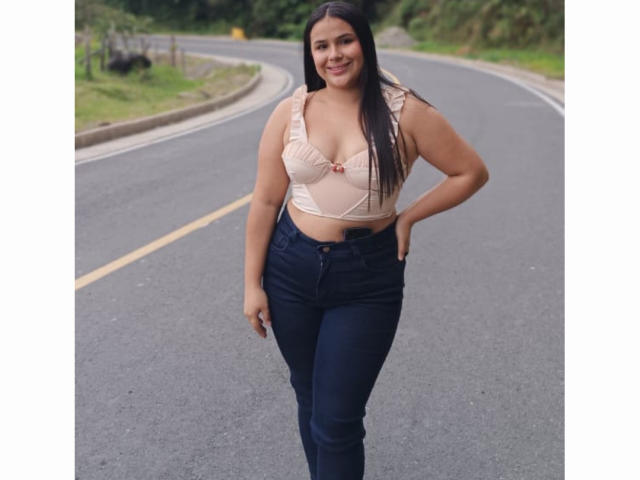 AmilyJones has medium tits, wearing a beige bra with pink bow, and dark blue jeans, standing on a curving road, smiling with long black hair, hands on hips.