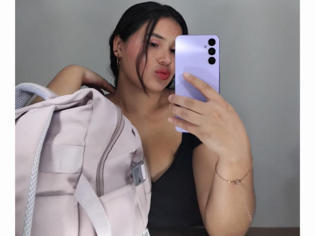 AmilyJones with dark hair, pink lipstick, black top, holding pink backpack, and purple phone, taking mirror selfie, showing her tits, wearing gold earrings, and delicate bracelet.