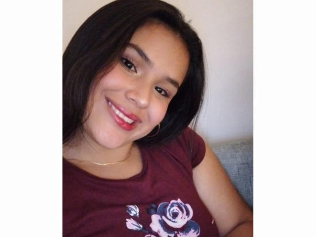 AmilyJones has medium brown skin, long black hair, and a cute smile with red lipstick. She's wearing a maroon shirt with a rose design, and hoop earrings.