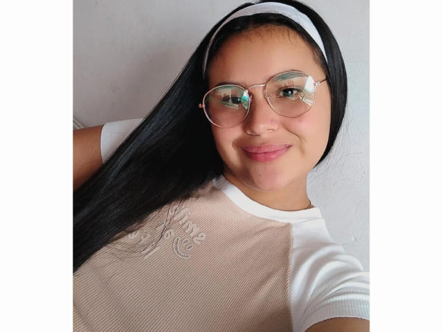 AmilyJones with long black hair, wearing a white headband, glasses, and a white shirt with embroidered details, smiling at the camera, showing her cute face and small nose.