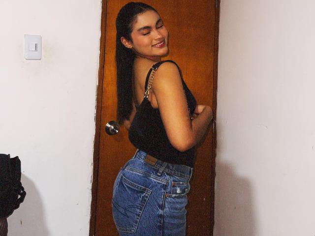MiiaPalmer with dark ponytail, smiling, wearing black tank top and high-waisted blue jeans, standing against wooden door, showing her ass and small tits.