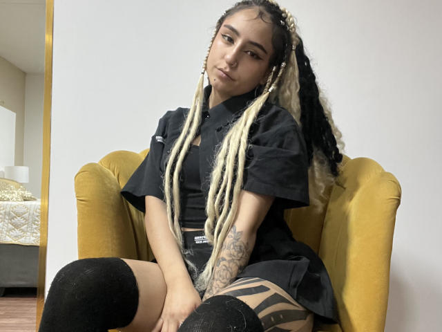 MaveParker with long blonde and black braided hair, black shirt, and black thigh-high socks, sitting on a yellow chair, showing tattooed arm and black shorts.