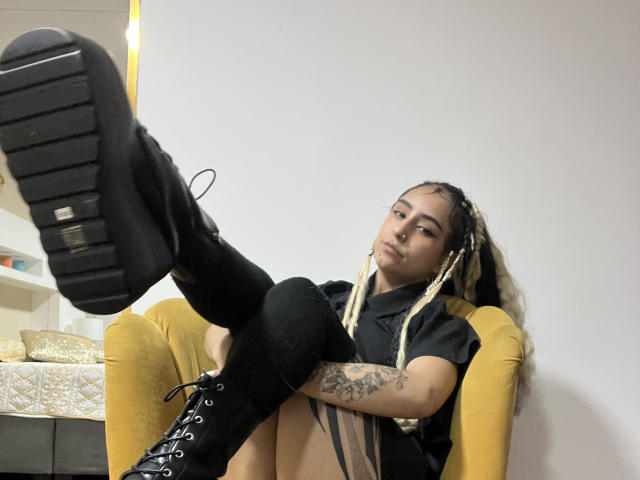 MaveParker with black boots, knee socks, and braided hair, showing her boot sole, tattoo on arm, and a confident expression, sitting in a yellow chair.
