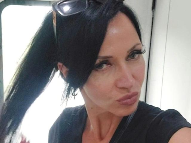 MiaAmour has puffy lips, dark straight ponytail, black shirt, sunglasses on head, and serious expression. She's a busty white woman with light skin and green eyes.