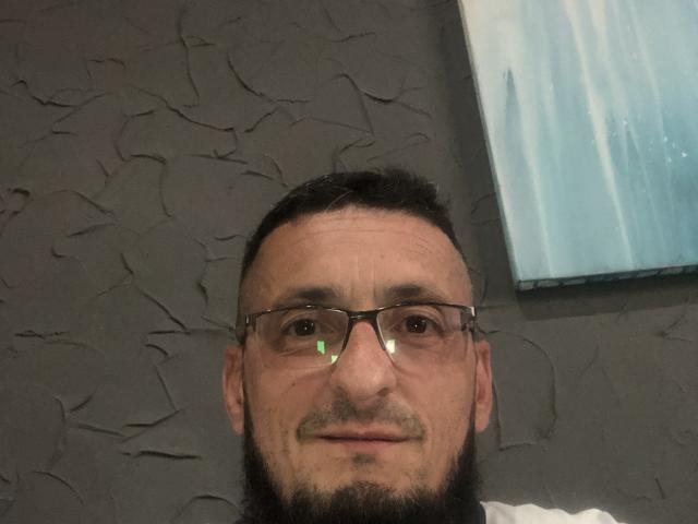 BernardFunny has a medium build, short black beard, glasses, and a slight smile. He's wearing a white shirt, with a textured gray wall behind him.