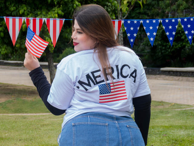 EymiTomsom with long brown hair, white shirt, black sleeves, blue jeans, holding tiny American flag, back to camera, outdoors, red-white-blue bunting, smiling, butt visible.