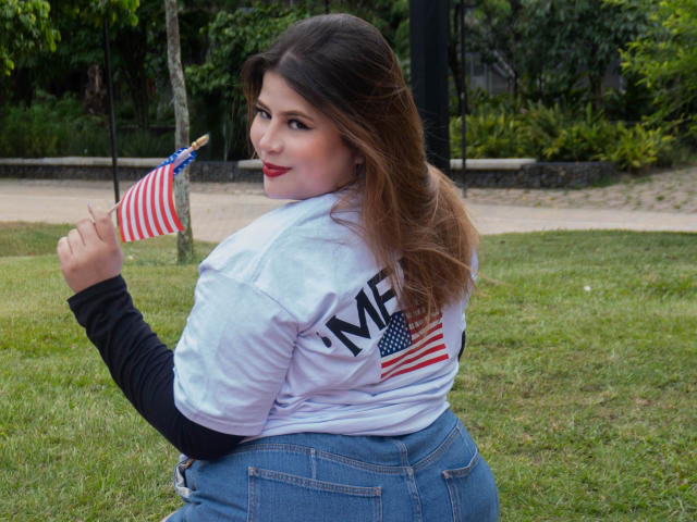 EymiTomsom, with red lipstick, holding a small American flag, wearing a white NY shirt, black sleeves, and blue jeans, sits on grass, looking back, with brown hair.
