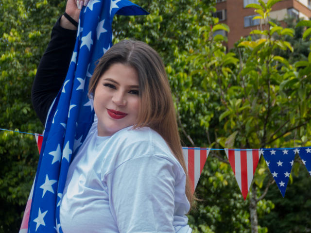 EymiTomsom with red lipstick, holding American flag, white shirt, long brown hair, smiling, green trees, red and white bunting, black sleeve, outdoors, summer, looking over shoulder.