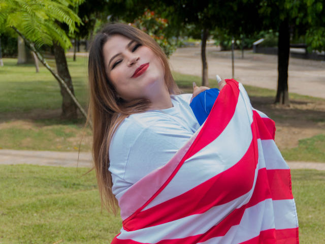 EymiTomsom with long brown hair, red lipstick, and a confident smile, wrapped in a red and white striped flag, holding a blue ball, standing in a grassy park.