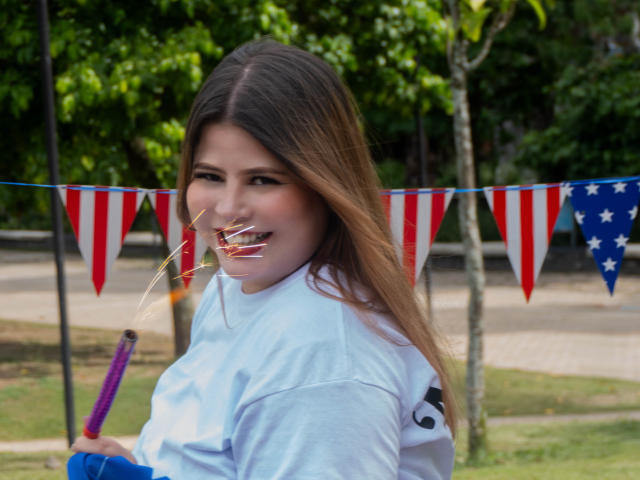 EymiTomsom with long brown hair, blue shirt, holding purple dildo in mouth, American flag bunting in background, grassy park, smiling, looking over shoulder.