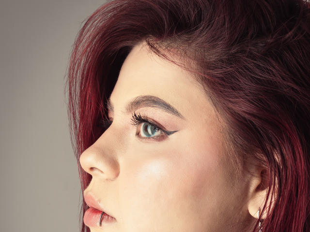 MelanieGate's profile shows a pale-skinned face with red hair, blue eyes, and winged eyeliner. Her nose is small, and her lips are slightly parted. Her tits are not visible.